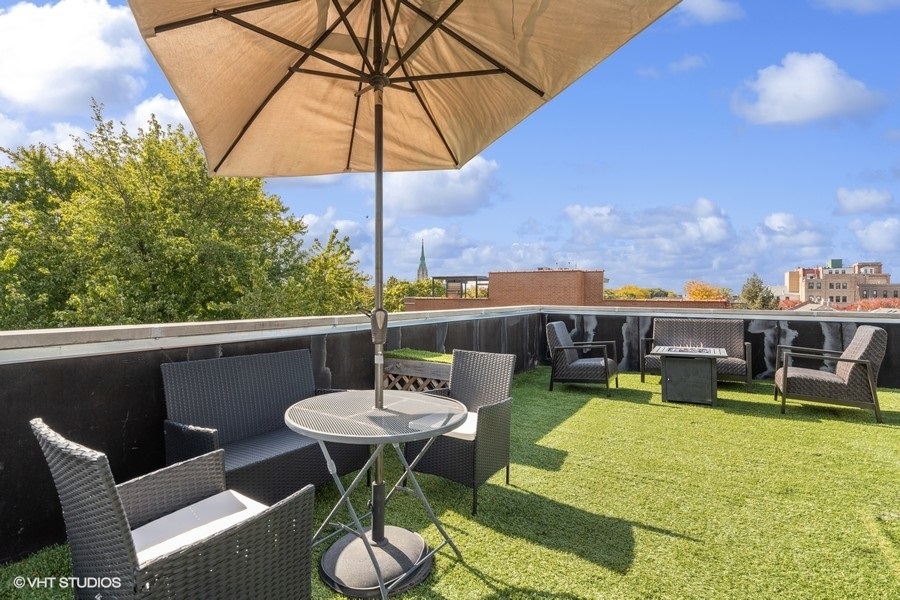 Rooftop patio with woven seating, artificial grass and a large umbrella overlooking nearby buildings.