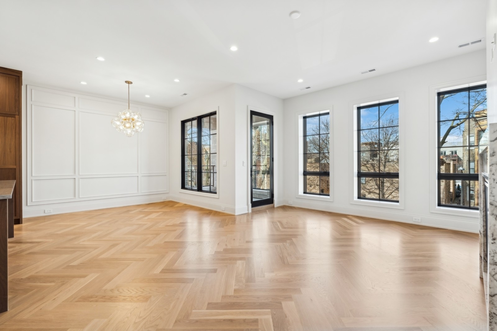 Empty living and dining area with herringbone floors, paneled walls, a chandelier, and tall black-framed windows.