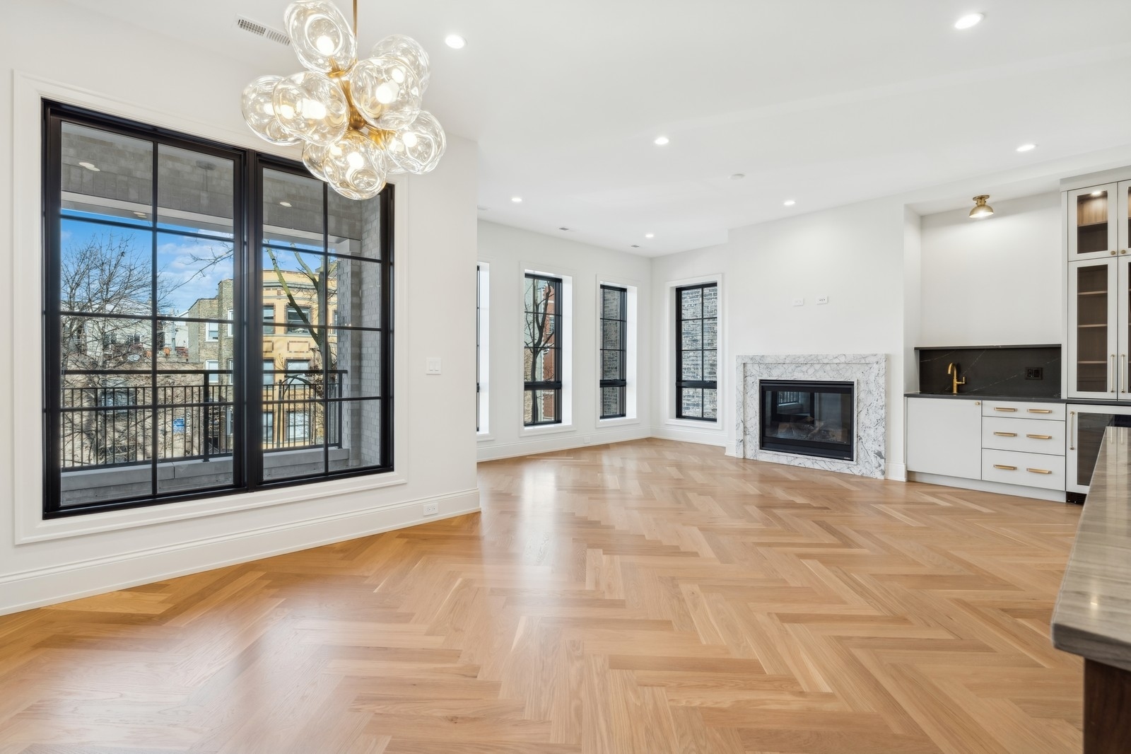Bright vacant living room with herringbone floors, marble fireplace, black-framed windows, and open kitchen.