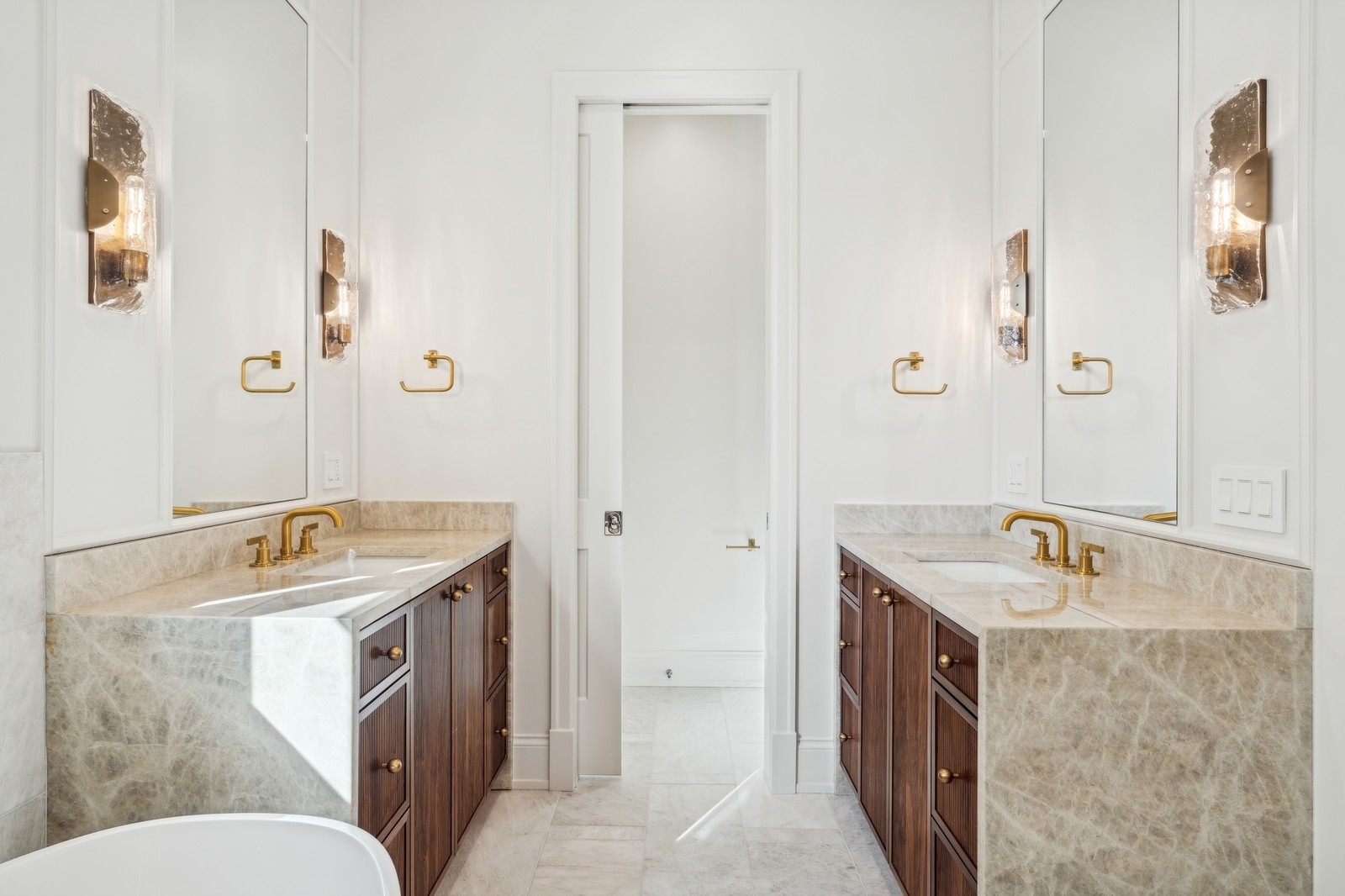 Bathroom with twin dark wood vanities, pale stone counters, brass fixtures, and a partially visible bathtub.