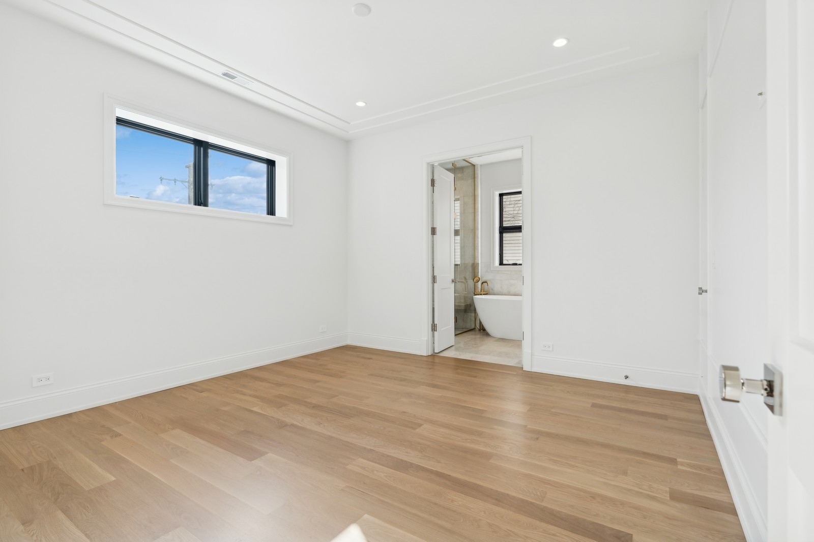 Empty bedroom with light oak floors and a doorway to an en-suite bathroom with a freestanding tub.