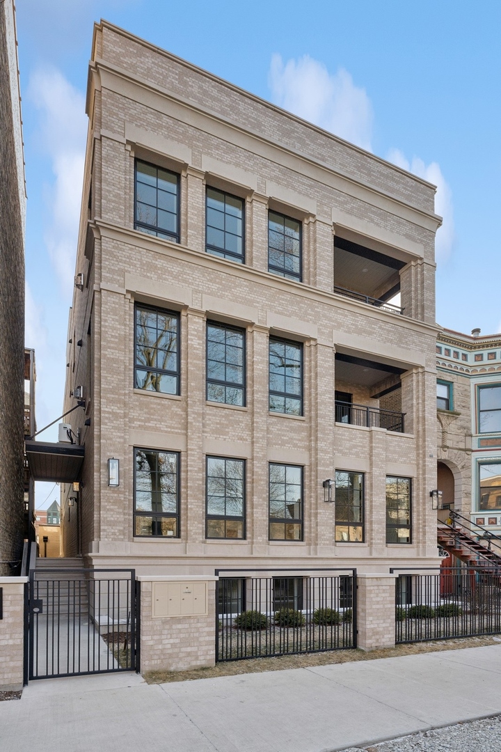 Light brick residential building with tall windows, recessed balconies, and a black metal sidewalk fence.