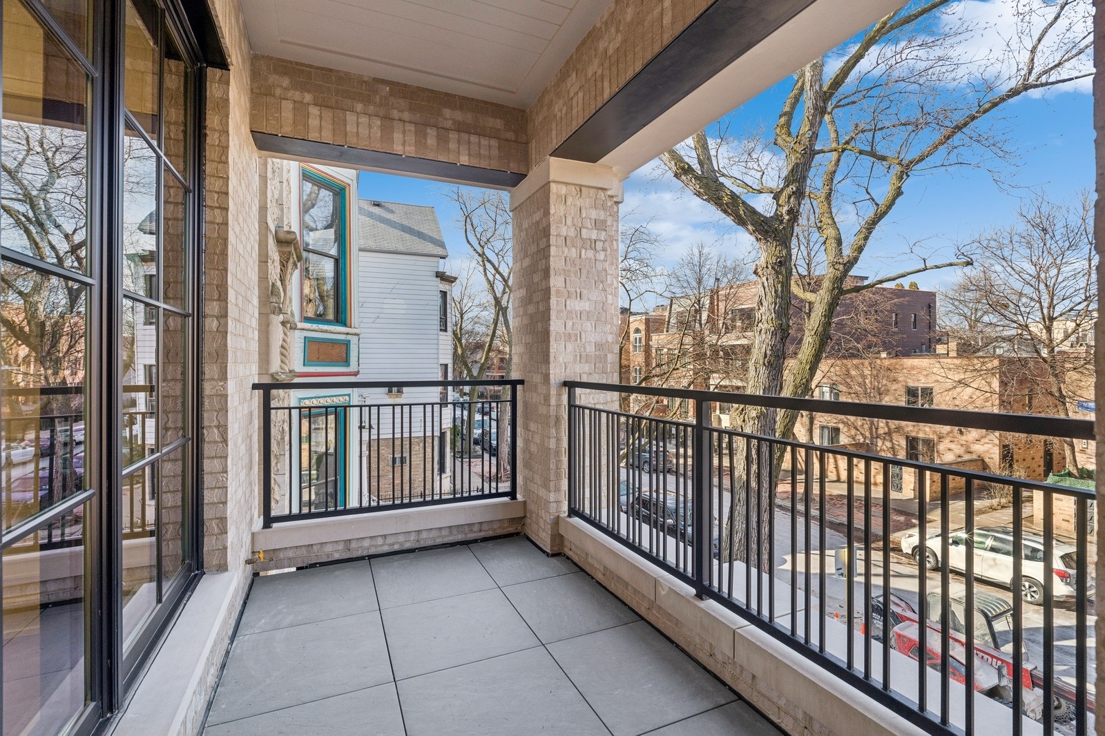 Covered balcony with gray tile, brick columns, black railings, and a view of the tree-lined street.