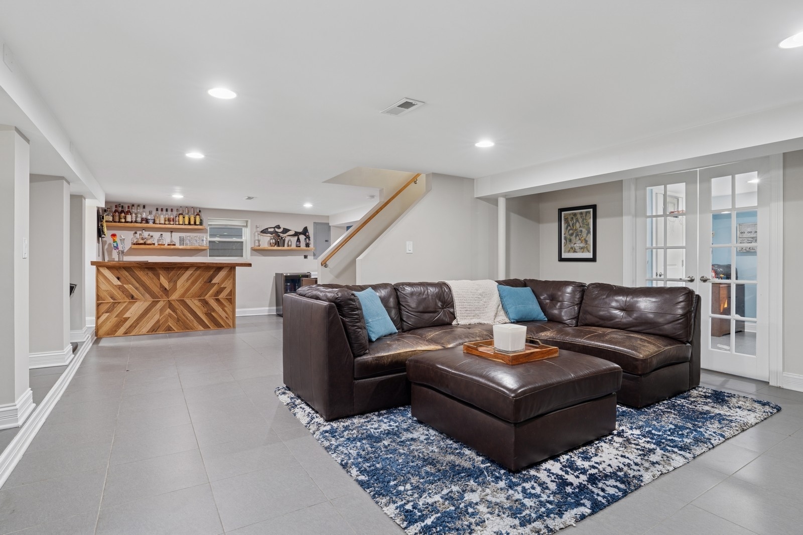 Basement living room with a leather sectional, blue-and-white rug, and wood-front bar.