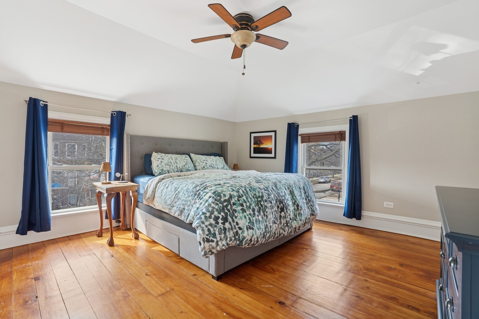 Bedroom with a patterned bed, blue curtains, and wide-plank hardwood floors.
