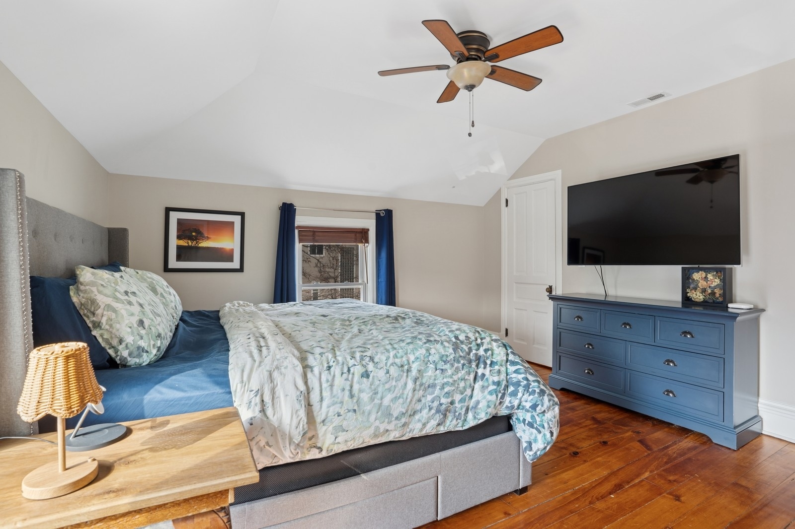 Furnished bedroom with a sloped ceiling, blue dresser and television, and polished hardwood floors.