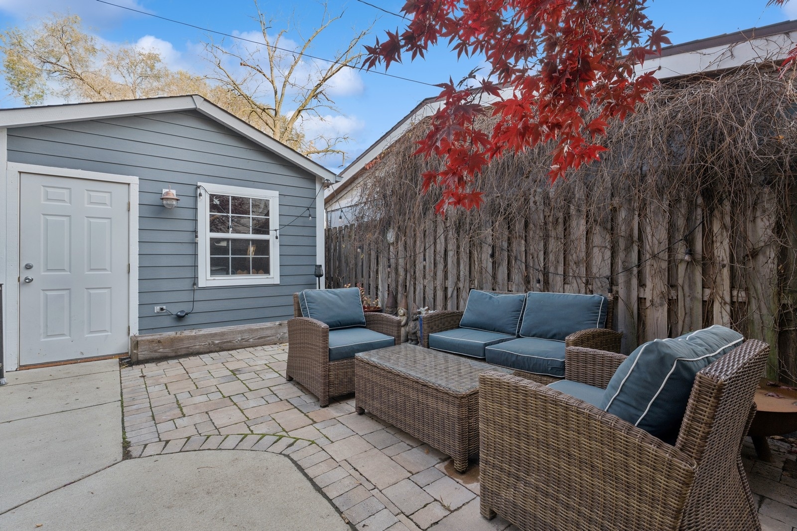 Furnished brick-paver patio with wicker seating, beside a gray outbuilding and vine-covered fence.