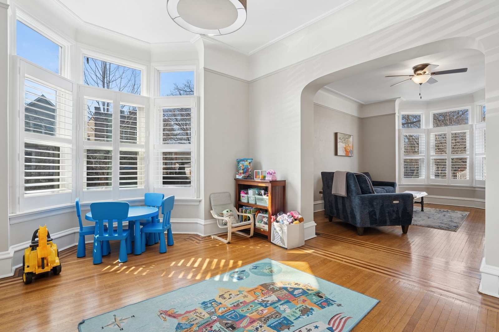 Bright play area with blue child-size furniture and toys beside a bay window, opening to a sitting room.