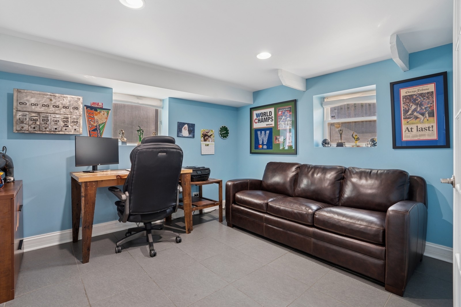 Home office with a wooden desk and chair beside a leather sofa, blue walls, and framed sports memorabilia.