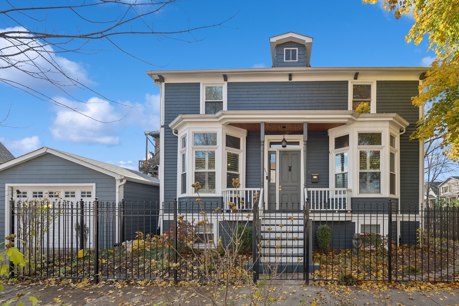 Blue-gray house with white-trimmed bay windows, covered porch, iron fence, and detached garage.