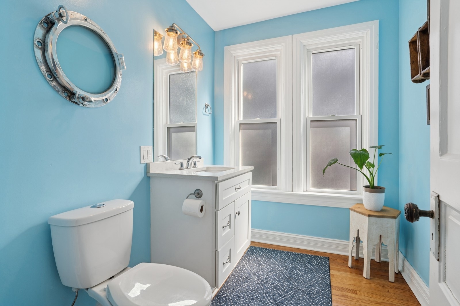 Bright turquoise bathroom with a white vanity, tall windows, and a porthole-style mirror.