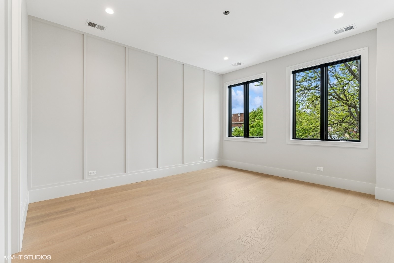 Empty room with pale hardwood floors, paneled walls, and two large windows overlooking trees.