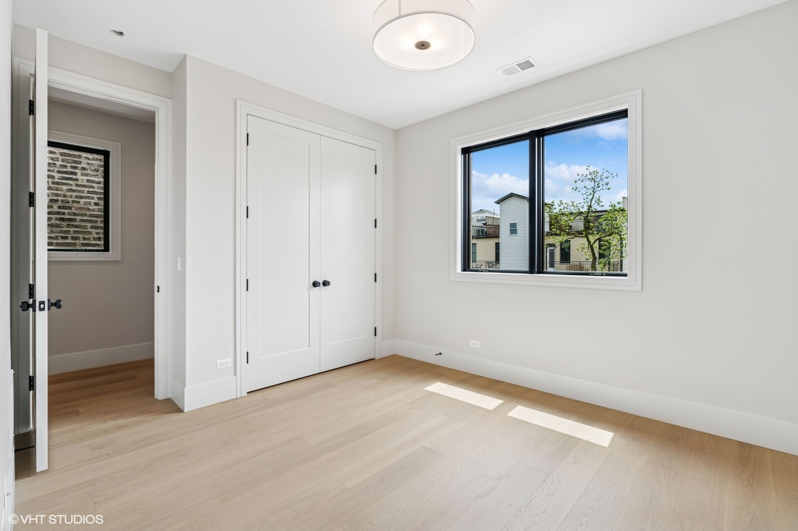 Vacant bedroom with pale wood floors, white closet doors, and a large window overlooking leafy trees.