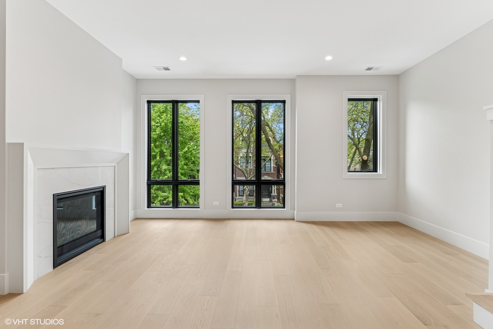 Vacant living room with a marble fireplace, pale hardwood floors, and tall black-framed windows.