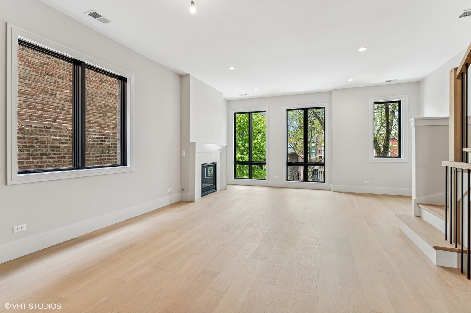 Empty living room with pale hardwood floors, a fireplace, tall windows, and a staircase.