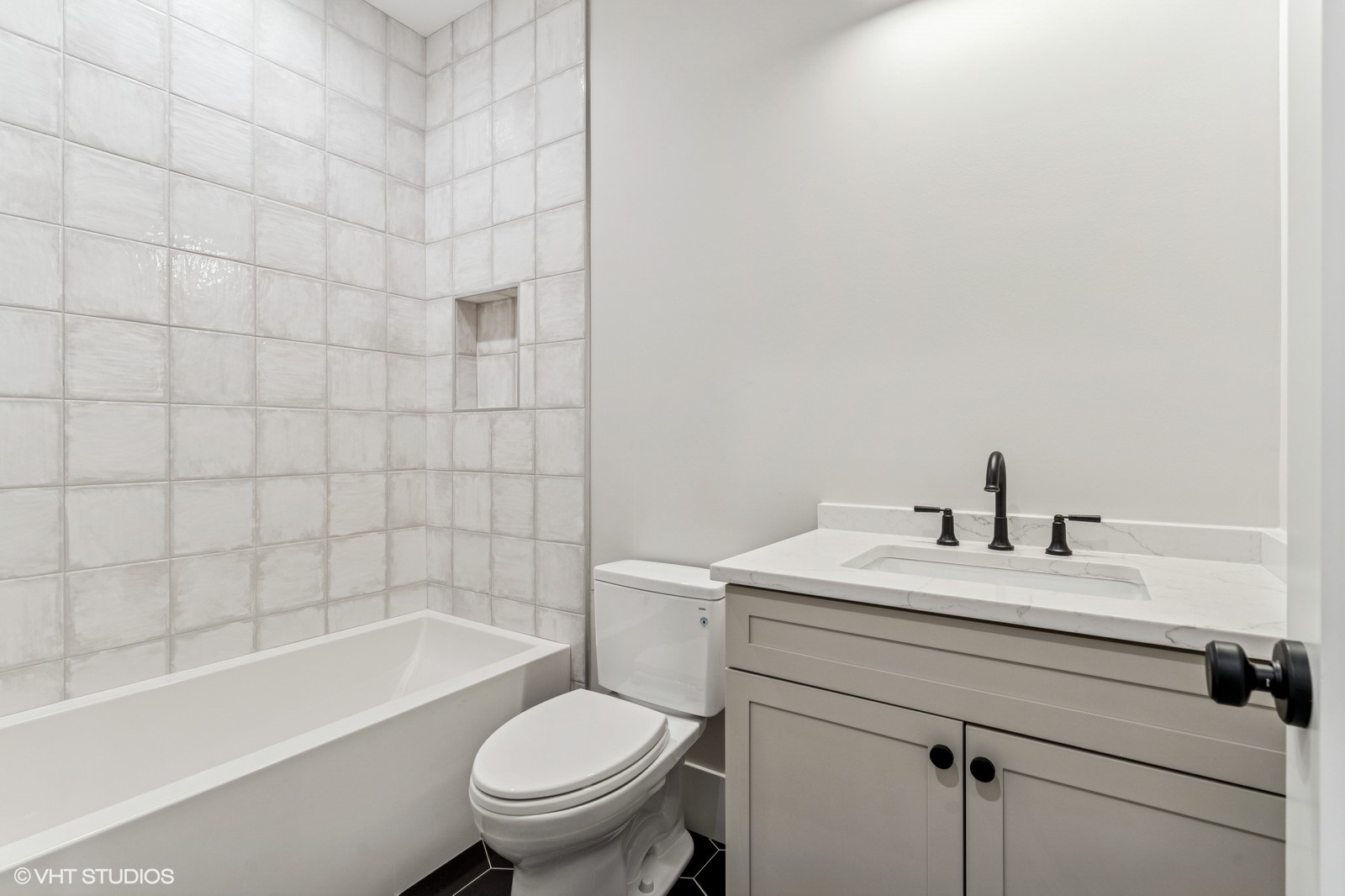 Bathroom with a tiled bathtub, recessed shower niche, toilet, and white vanity with black fixtures.