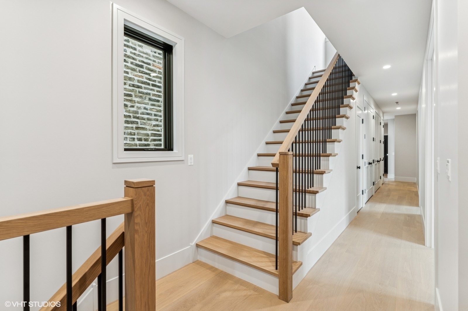 Light wood staircase with dark metal balusters beside a bright hallway and brick-facing window.