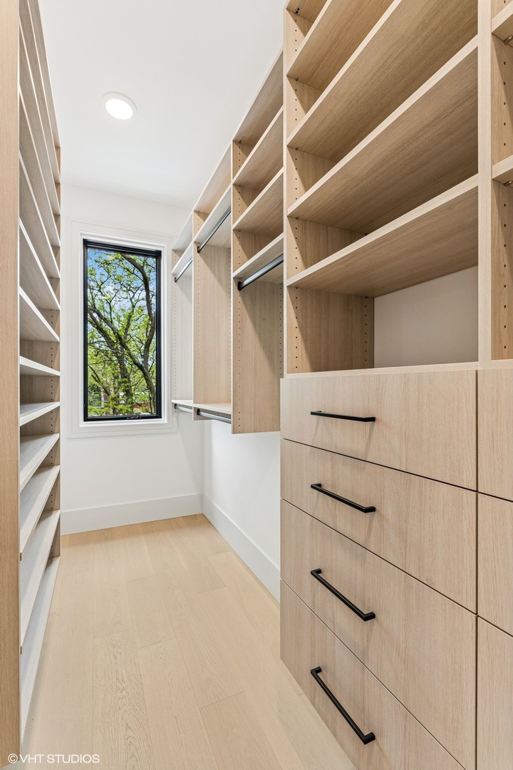 Walk-in closet with light wood shelving and drawers beside a tall window overlooking leafy trees.