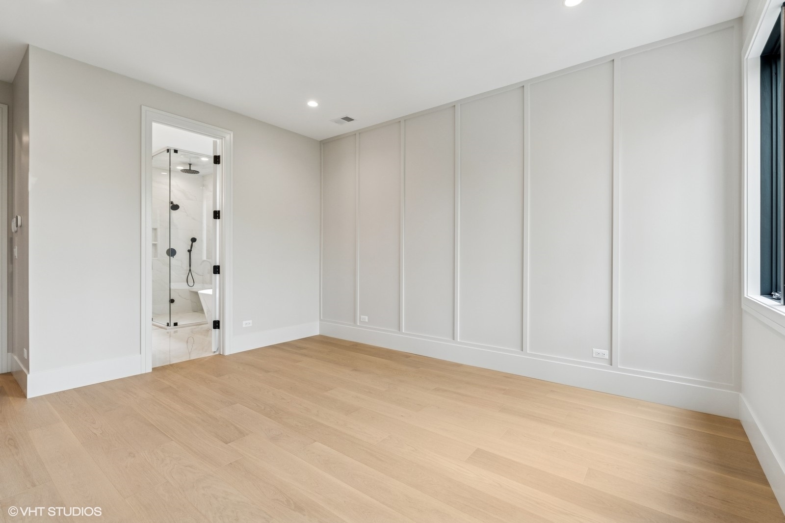Empty bedroom with light oak floors and paneled walls, opening to a bathroom with a glass shower.