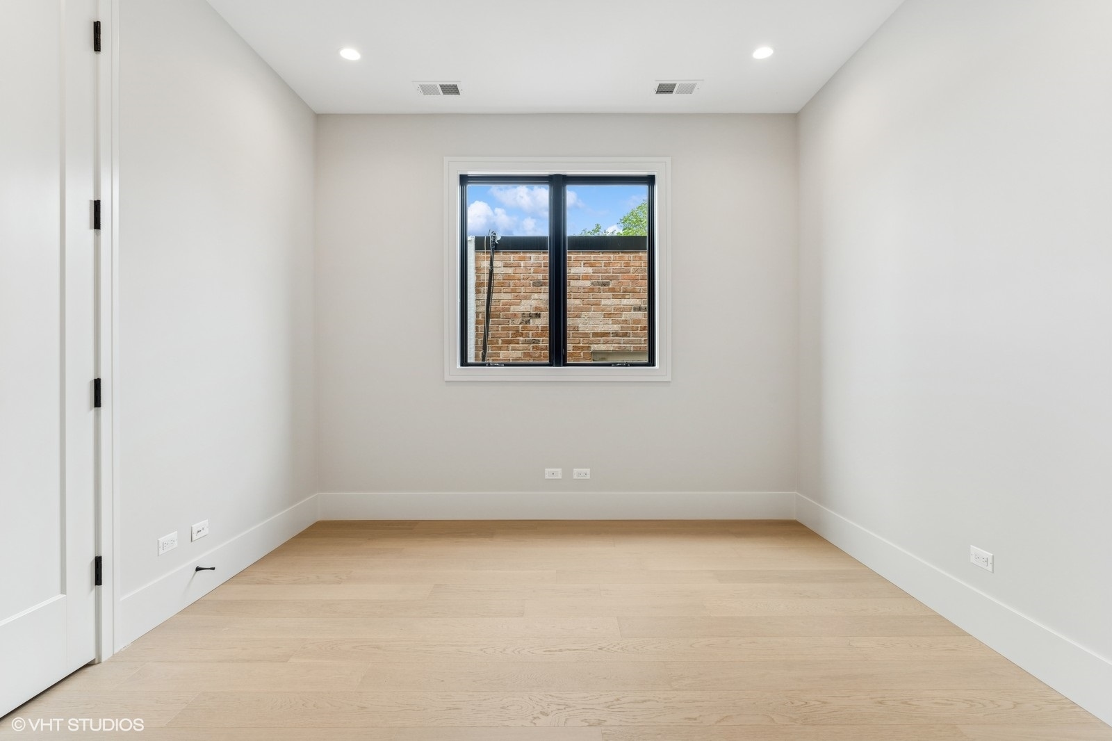 Empty room with pale hardwood floors, recessed lights, and a black-framed window overlooking a brick wall.
