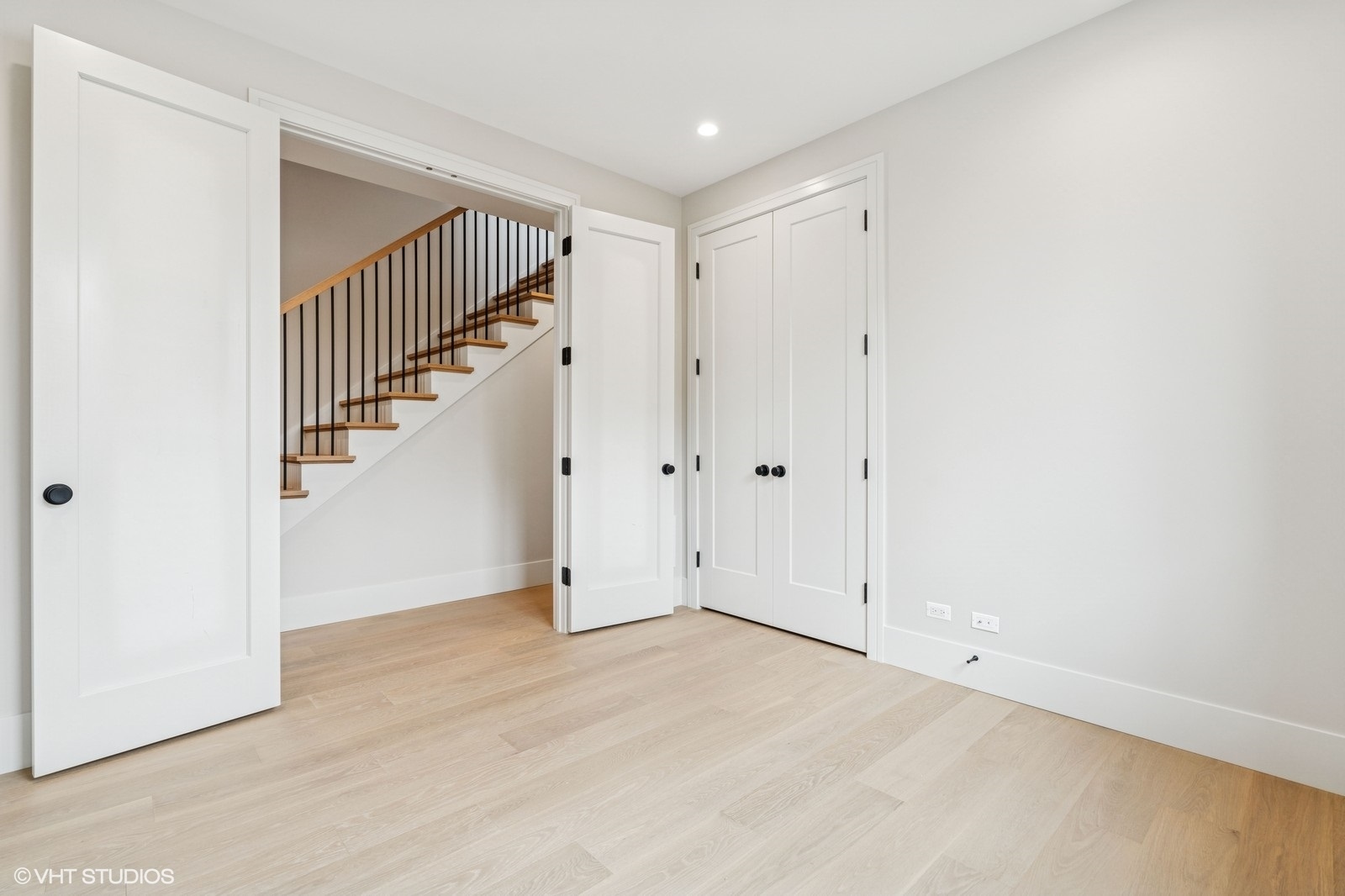 Bright empty room with pale wood floors and an open doorway to a staircase with black balusters.