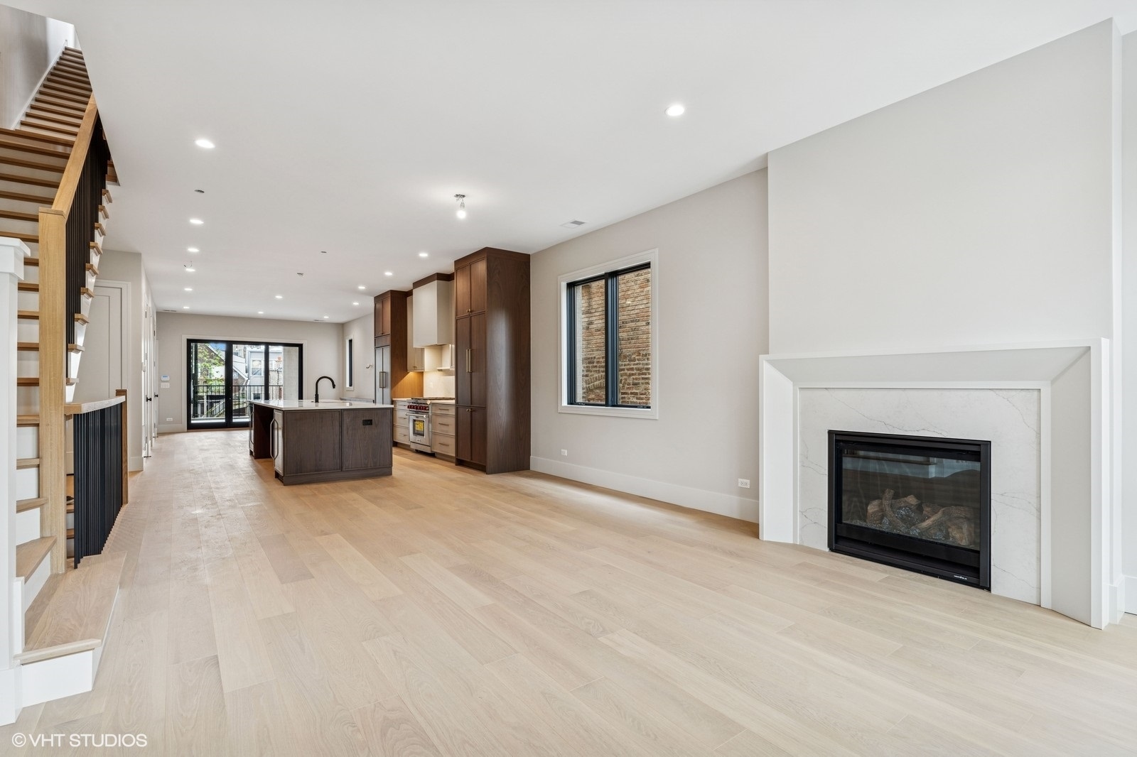 Open-plan living room with a marble-surround fireplace, dark kitchen cabinetry, and a wood staircase.