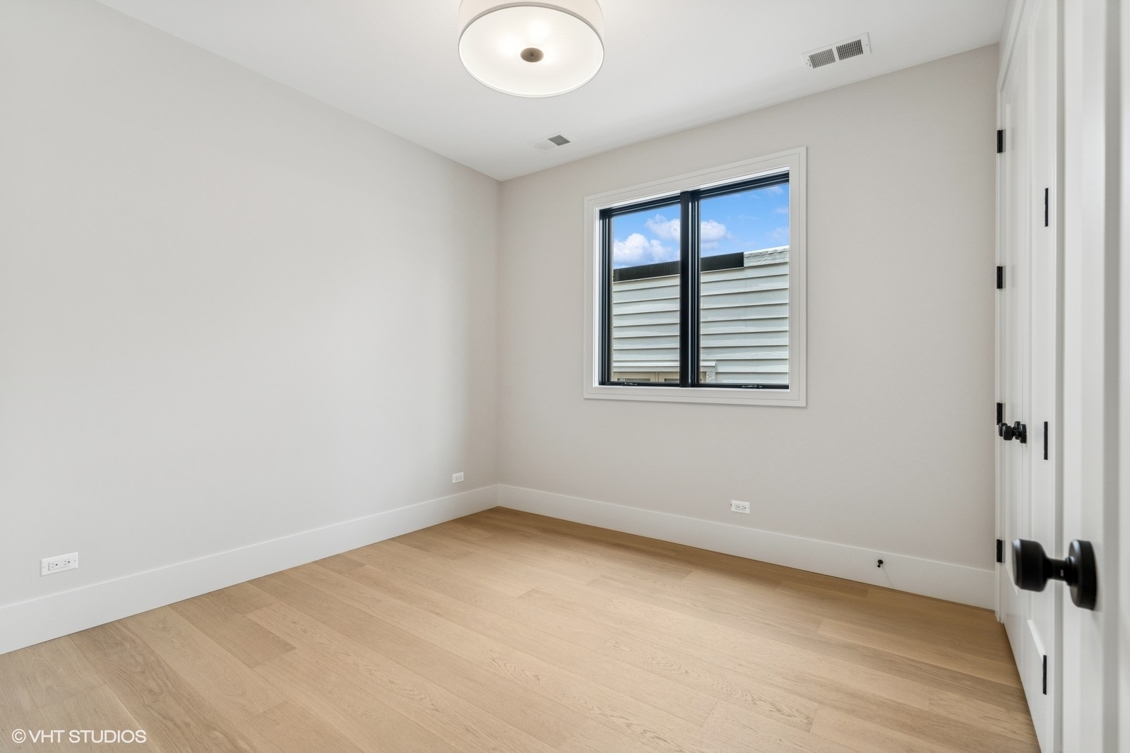 Empty bedroom with pale wood floors, a window, and white closet doors.