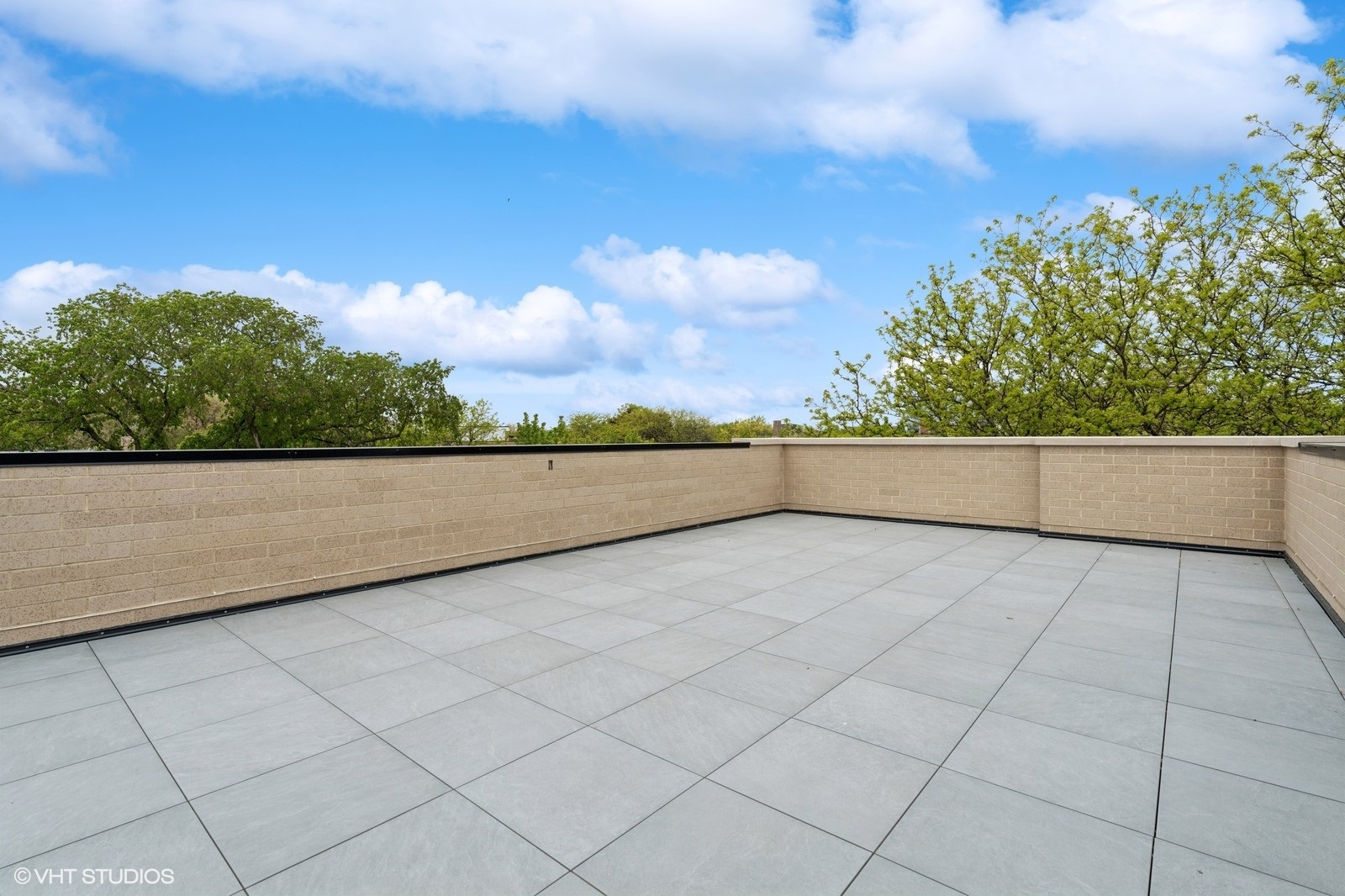 Rooftop terrace with gray pavers, tan masonry walls, and leafy trees beyond.