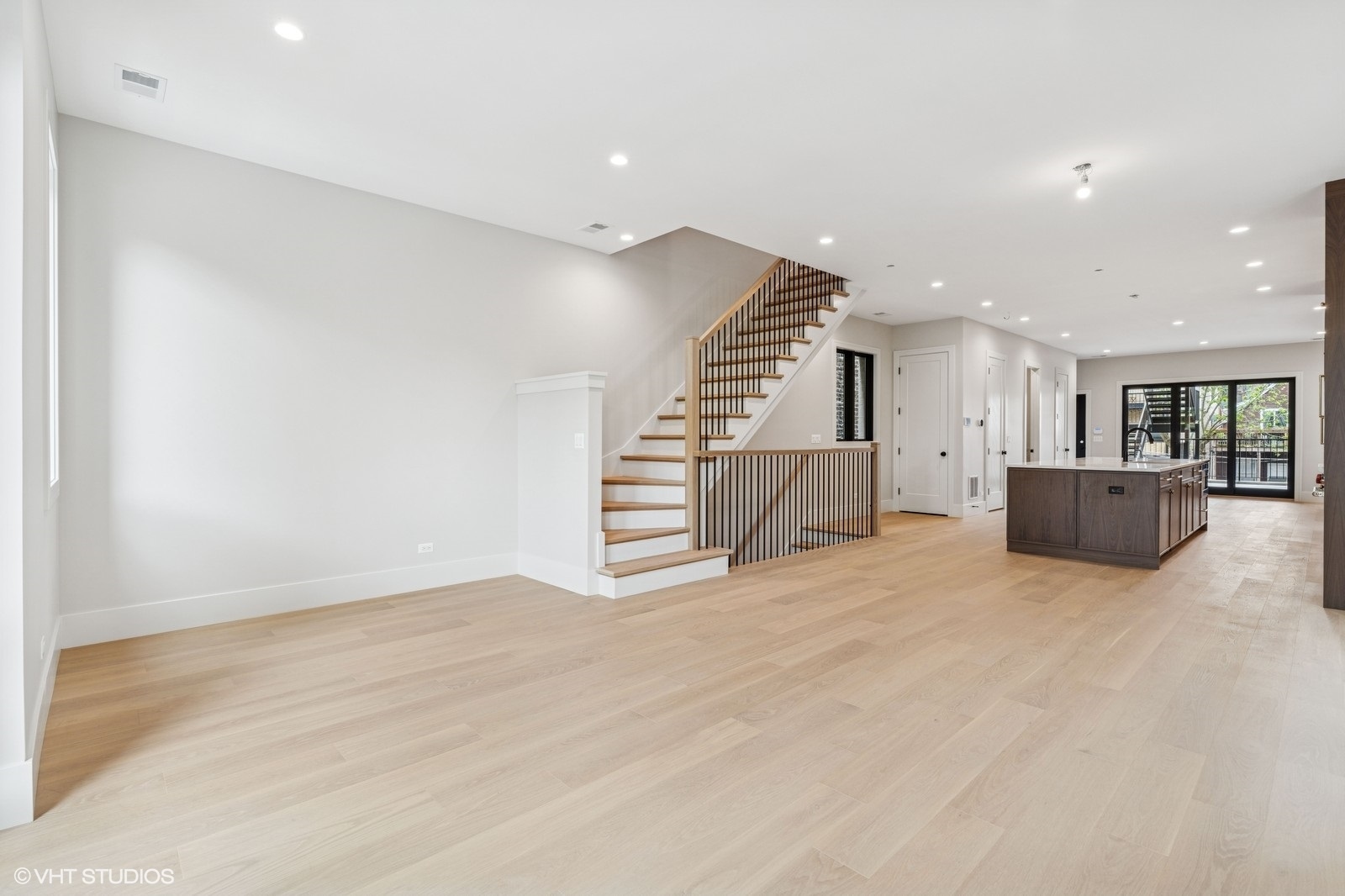 Open-plan living area with pale wood floors, a wood-and-metal staircase, and a dark kitchen island.