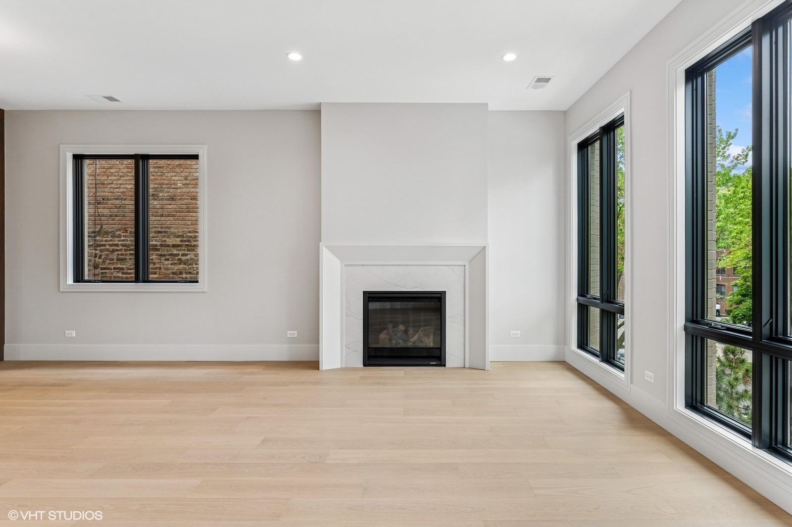 Unfurnished living room with a marble-surround fireplace, pale wood floors, and tall black-framed windows.