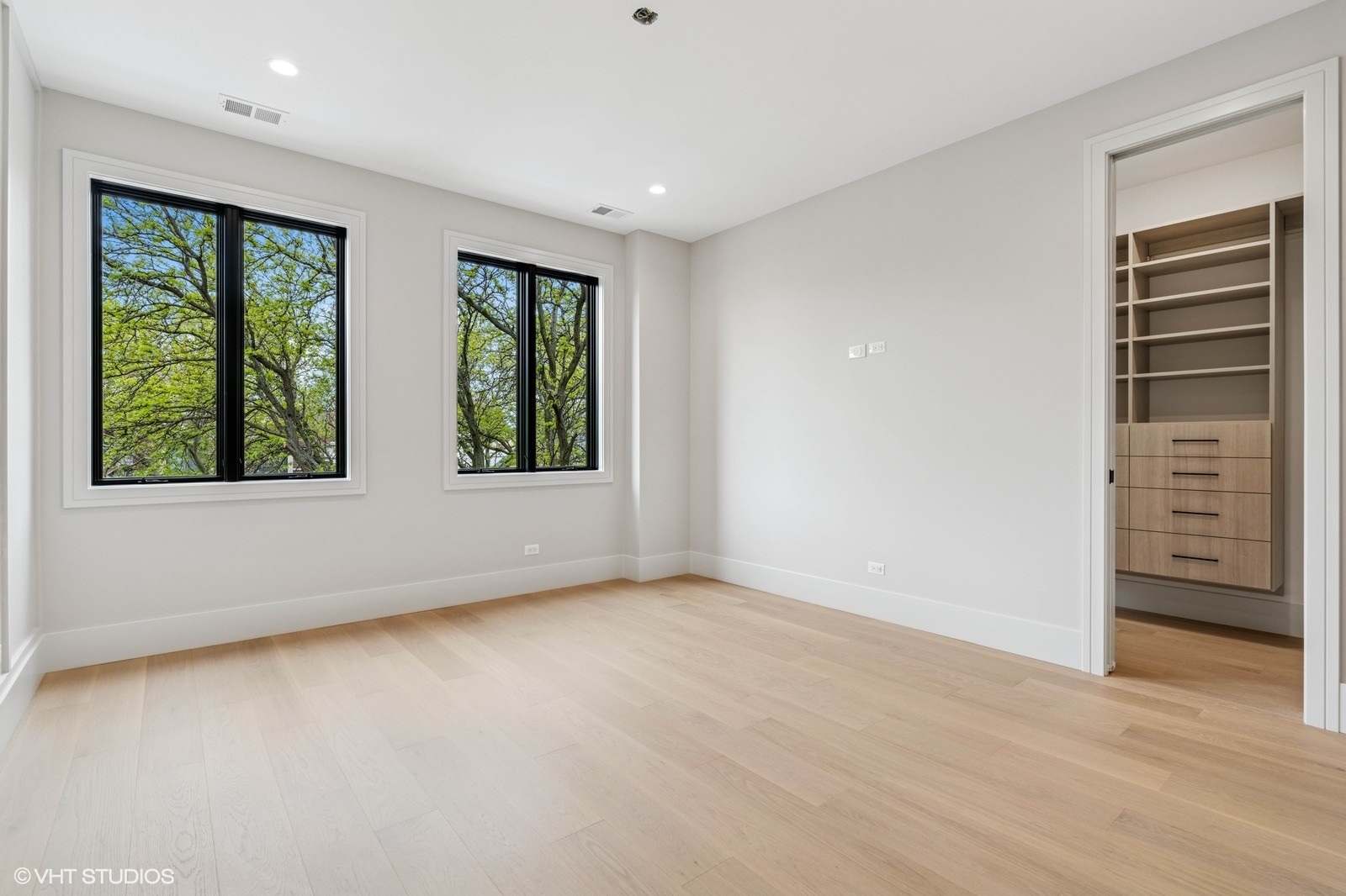 Empty bedroom with pale hardwood floors, two large windows, and a built-in closet.