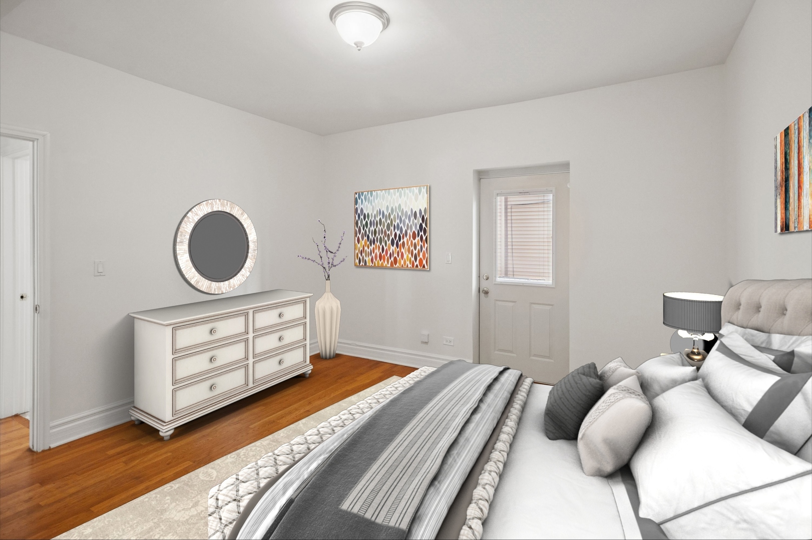 Furnished bedroom with gray-and-white bedding, hardwood floors, and a white dresser beneath a round mirror.