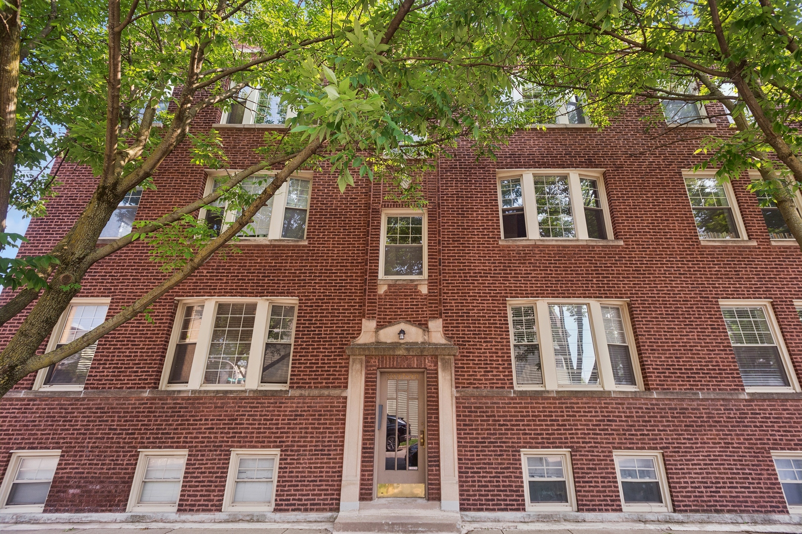 Red brick apartment building with a stone-trimmed entrance, framed by leafy trees.