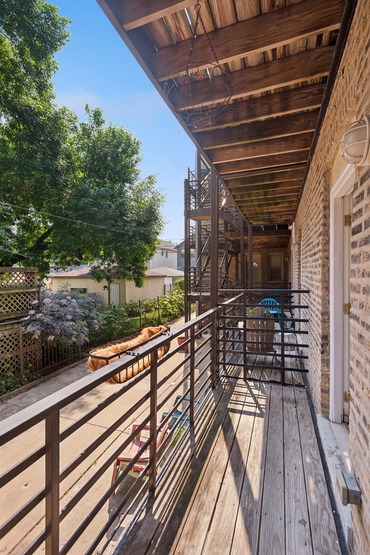 Wooden balcony with metal railings and overhead canopy beside a brick building, overlooking a leafy garden.