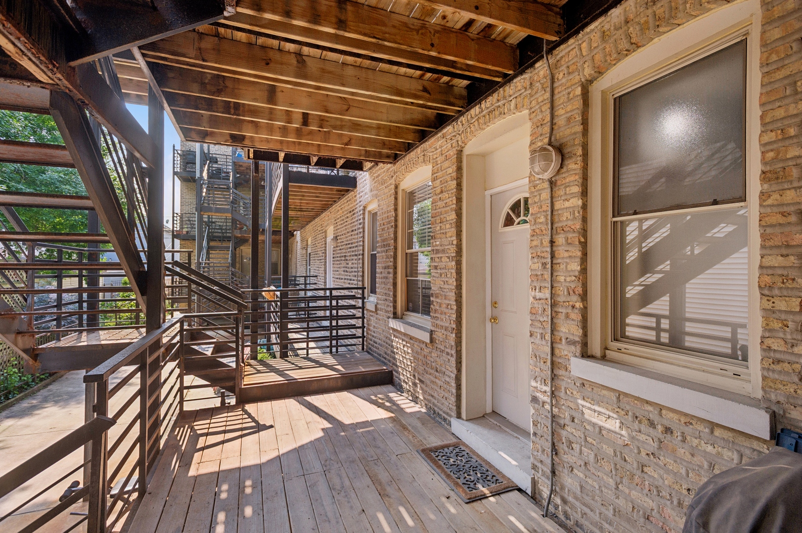 Covered wooden entry porch beside a brick apartment building, with dark railings, exposed beams, and a white door.