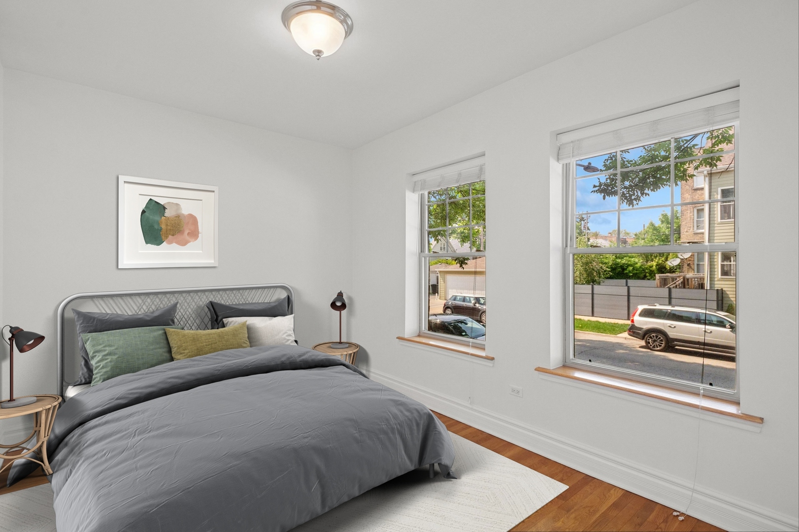 Staged bedroom with a gray bed, abstract wall art, and two large street-facing windows.