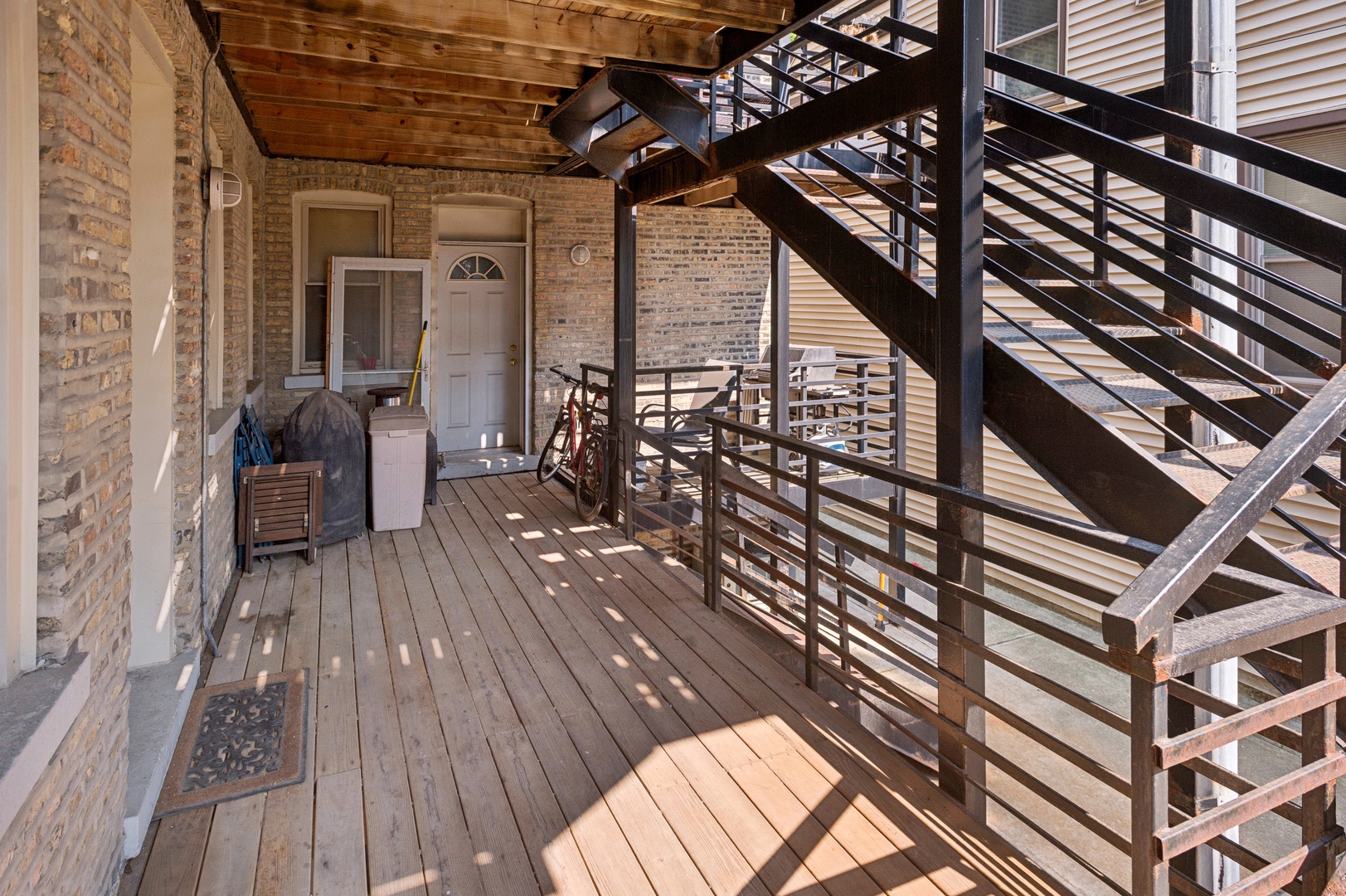 Covered wood entry deck with a brick wall, white door, and black metal stairs.