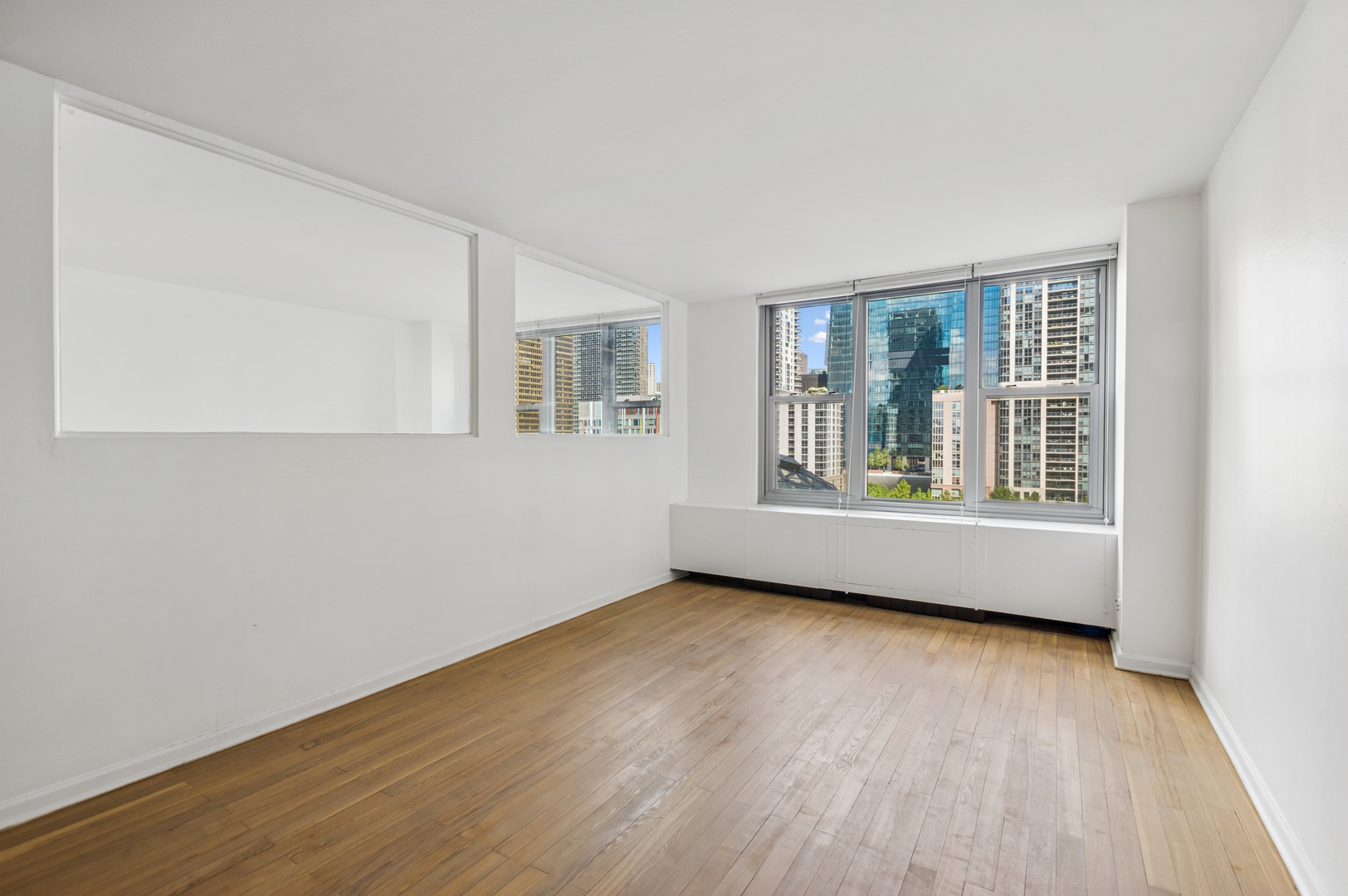 Vacant room with hardwood floors, a wall mirror, and large windows overlooking downtown buildings.