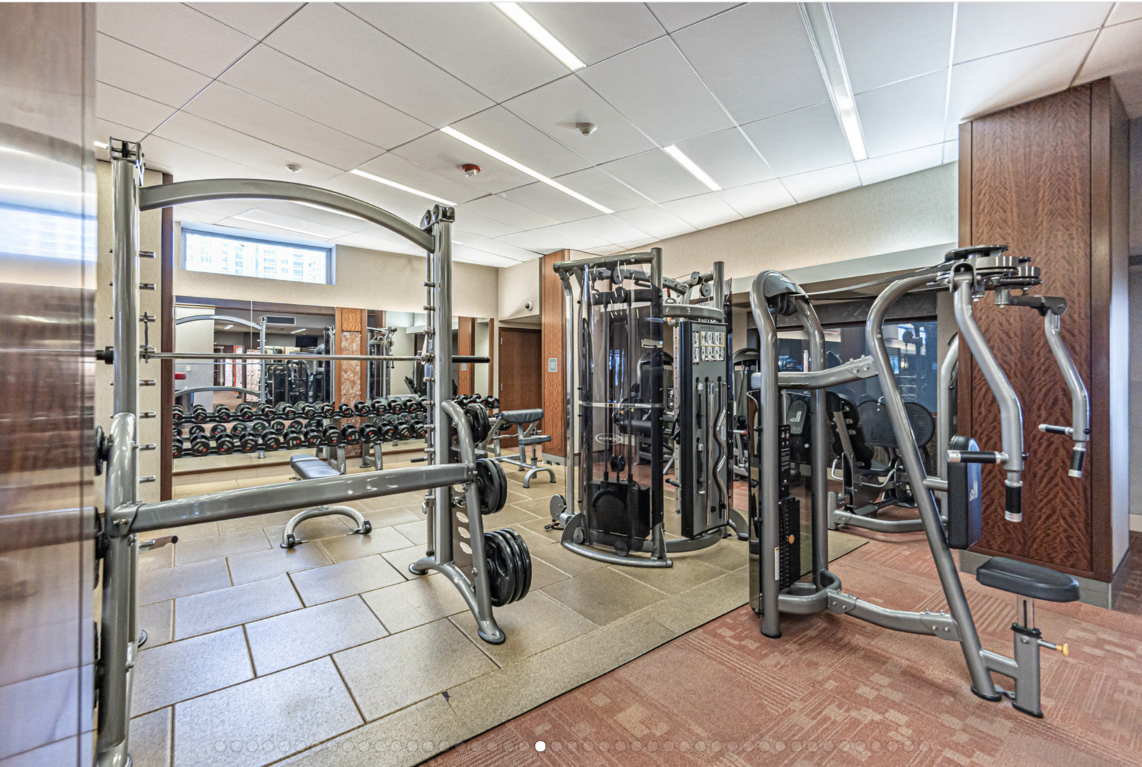 Building fitness center with weight machines, a squat rack, and dumbbells reflected in a wall mirror.