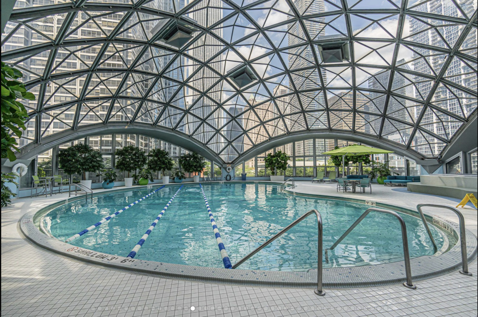 Indoor pool beneath a geometric glass dome, with lane dividers and city skyscrapers beyond the windows.
