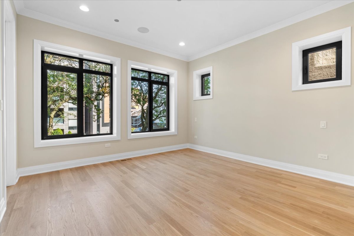Vacant room with light hardwood floors and several black-framed windows overlooking trees and nearby buildings.