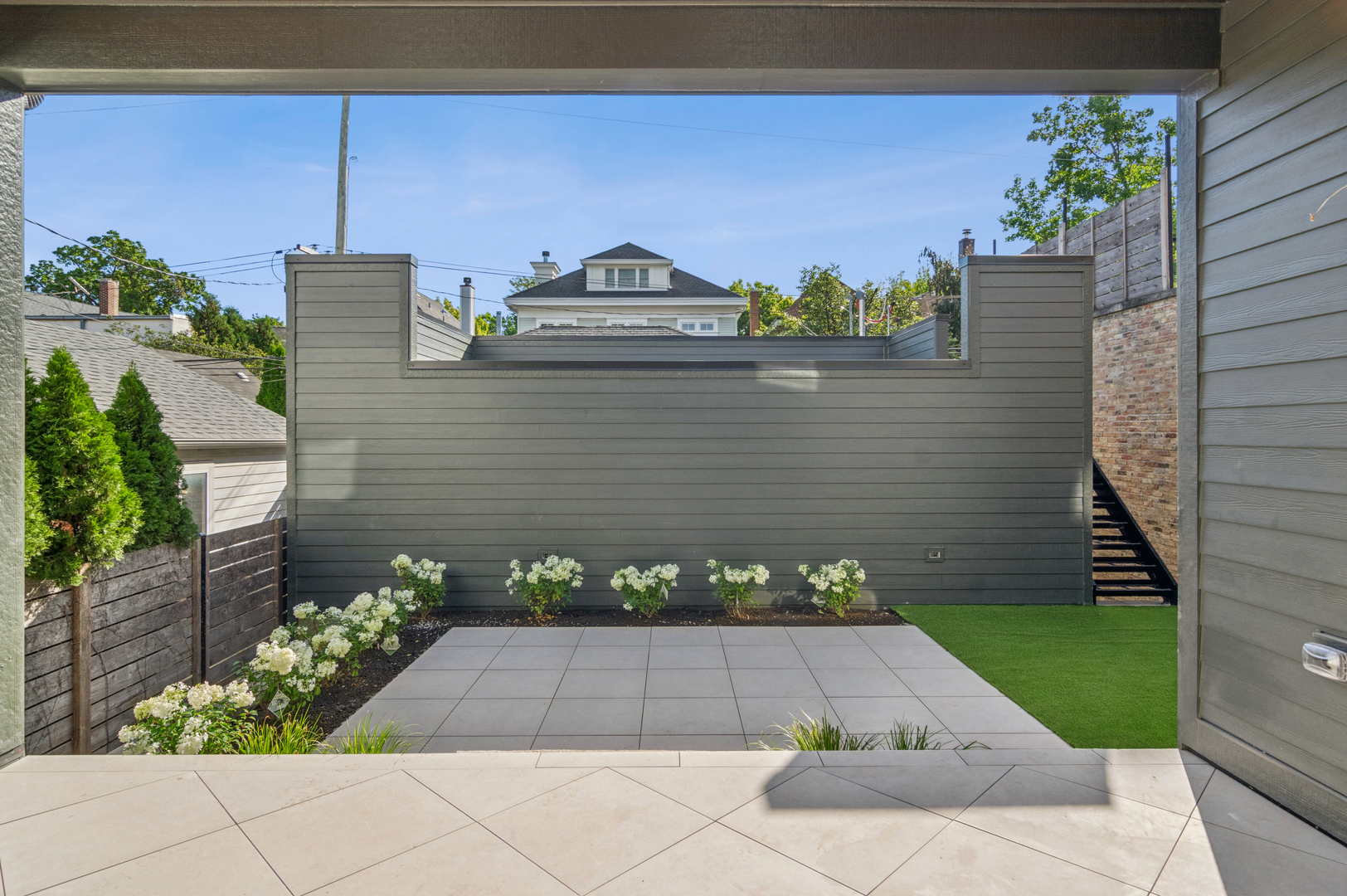 Outdoor patio with broad pavers, green turf, flower beds, and tall privacy fencing.