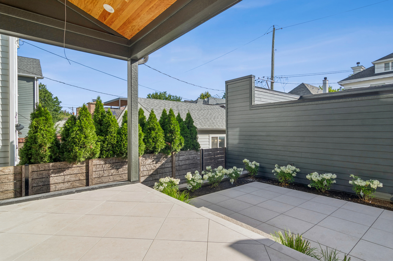 Tiled outdoor patio with a covered wood ceiling, gray privacy fence, and white flowering plants.