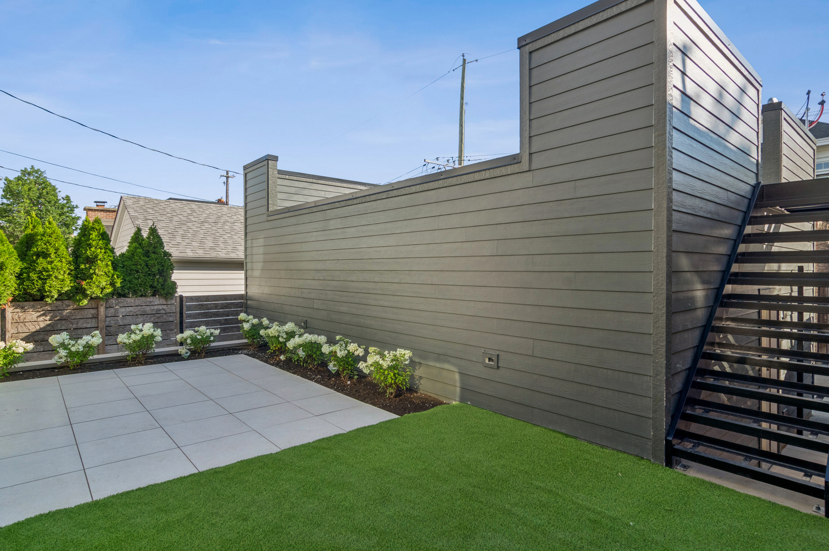 Outdoor patio with gray pavers, green lawn, planted shrubs, and an open staircase.