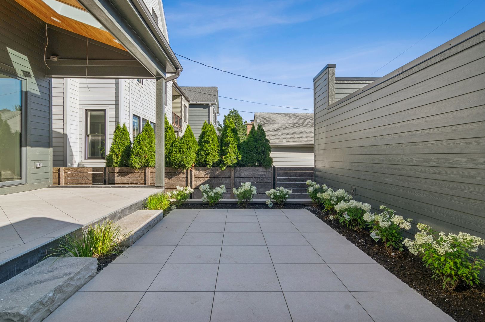 Paved patio beside a modern home, bordered by privacy fencing and white flowering plants.