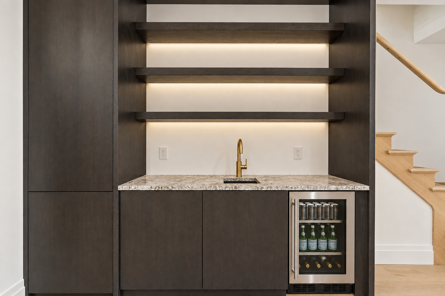 Wet bar with illuminated shelves, stone counter, brass faucet, and beverage refrigerator beside a staircase.