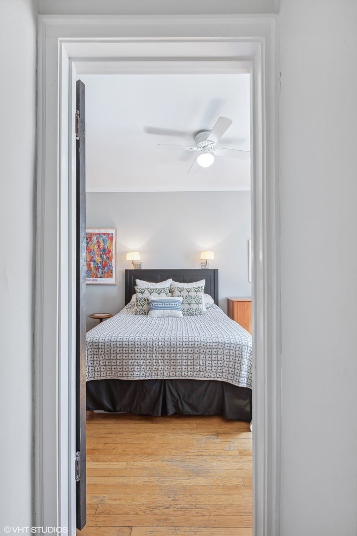Bedroom viewed through a doorway, with a patterned bedspread, dark headboard, and hardwood floor.