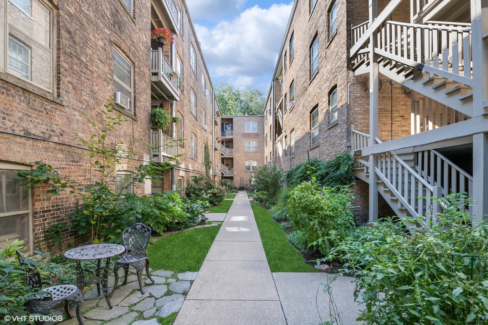 Apartment courtyard with a central walkway, lush garden beds, café seating, and white exterior staircases.