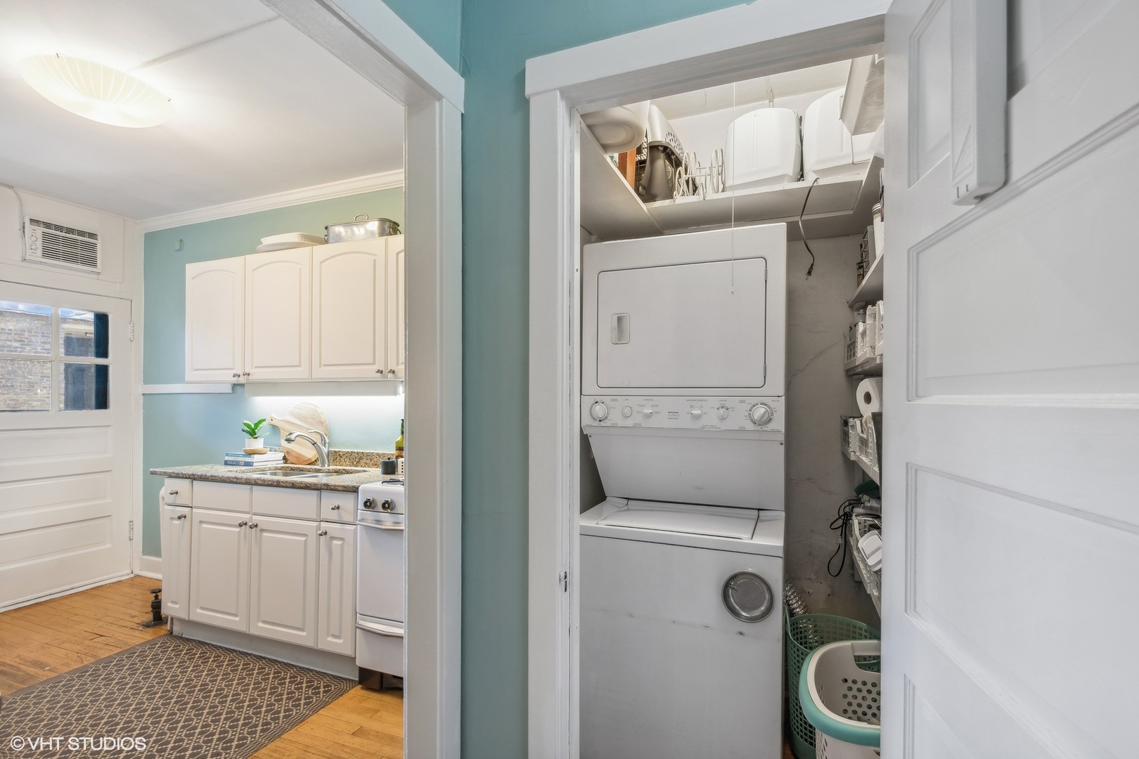 Laundry closet with stacked washer and dryer beside a compact kitchen with white cabinets and teal walls.