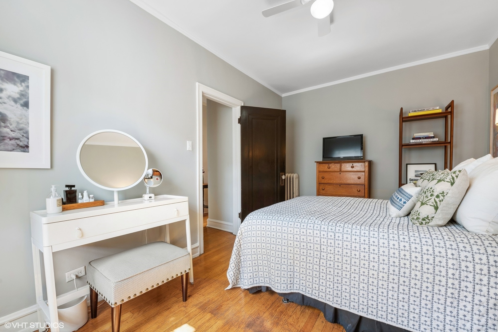 Bedroom with a patterned bed, white vanity and round mirror, and warm hardwood floors.